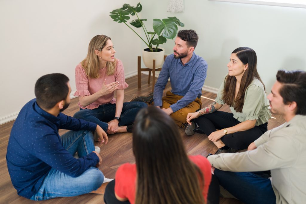 Groupe de personnes assises en cercle par terre, participant à un cercle de parole dans un espace collectif thérapeutique, échange bienveillant et partage d’émotions.